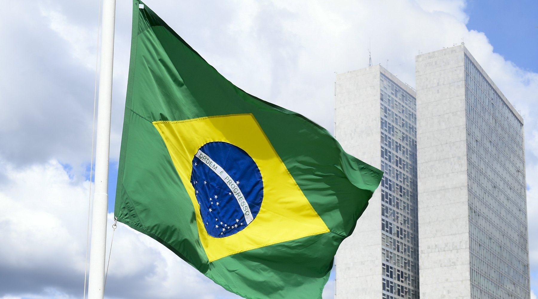 Bandeira do Brasil tremulando ao lado das duas torres do Congresso Nacional, em Brasília, com céu azul e nuvens ao fundo.